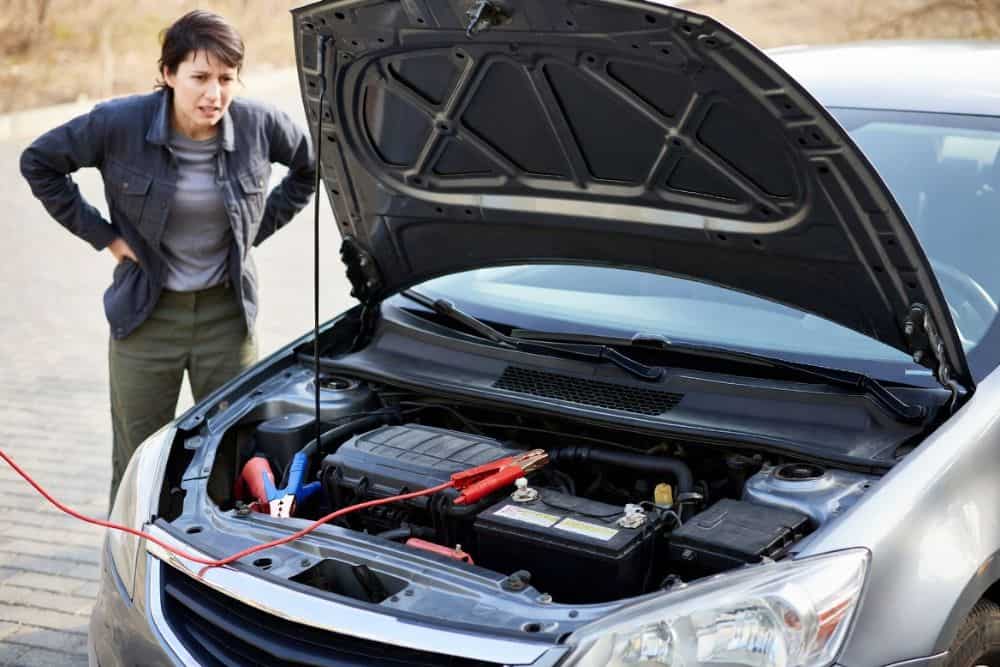 A person standing next to their car with a dying car battery, and a jumper cable laying across the engine
